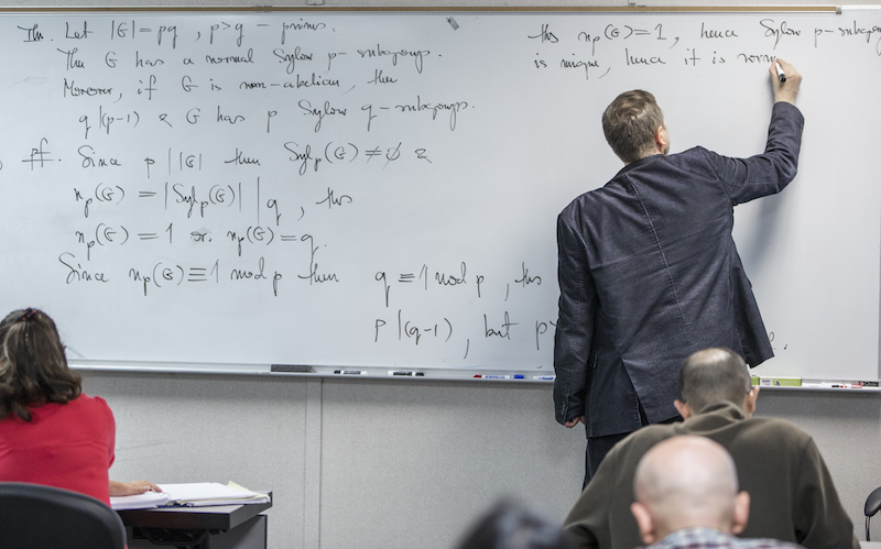 A professor writes an equation on a whiteboard in a classroom.